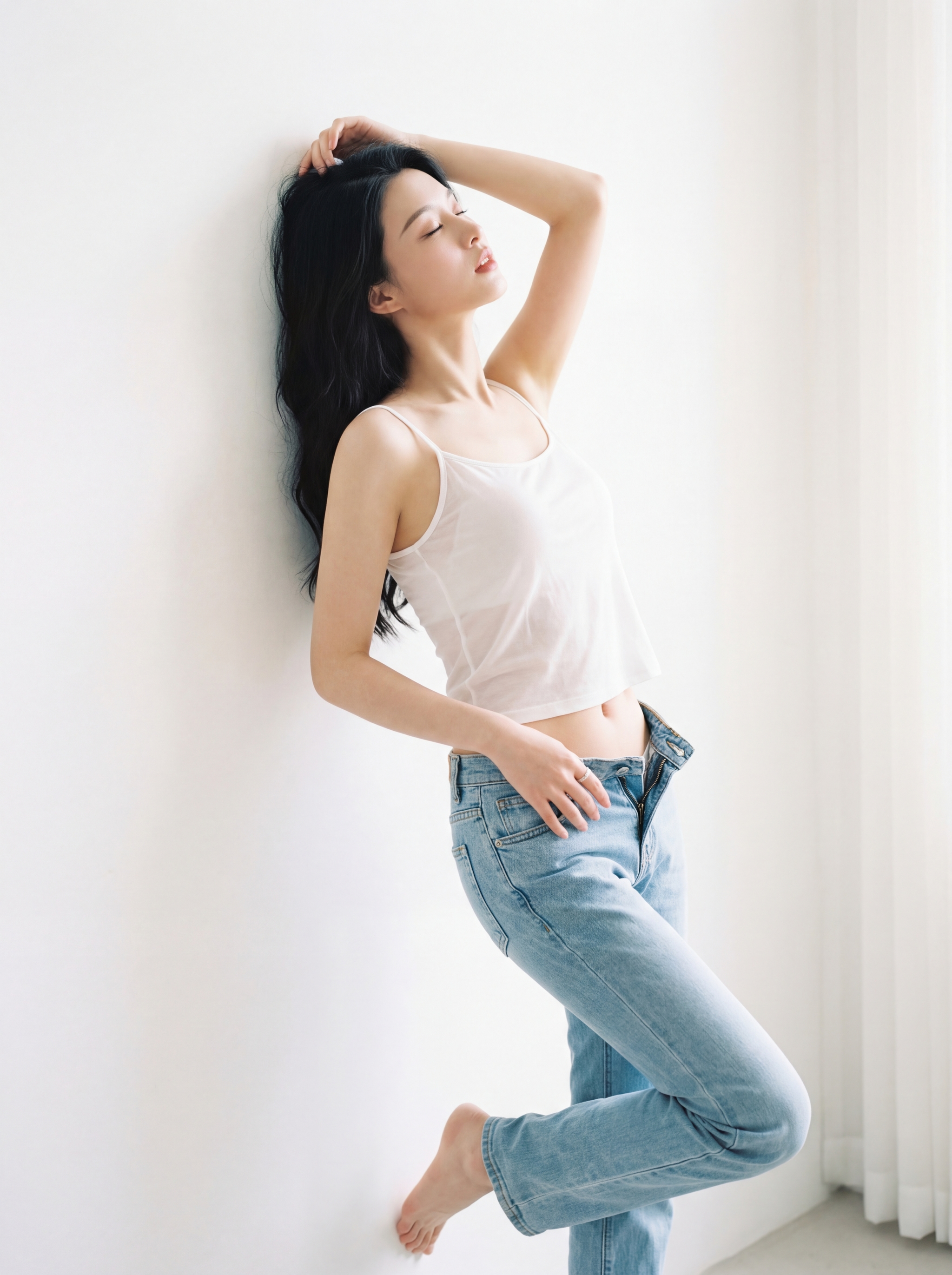 A person in a white tank top and blue jeans leaning against a wall near a bright window in soft natural light