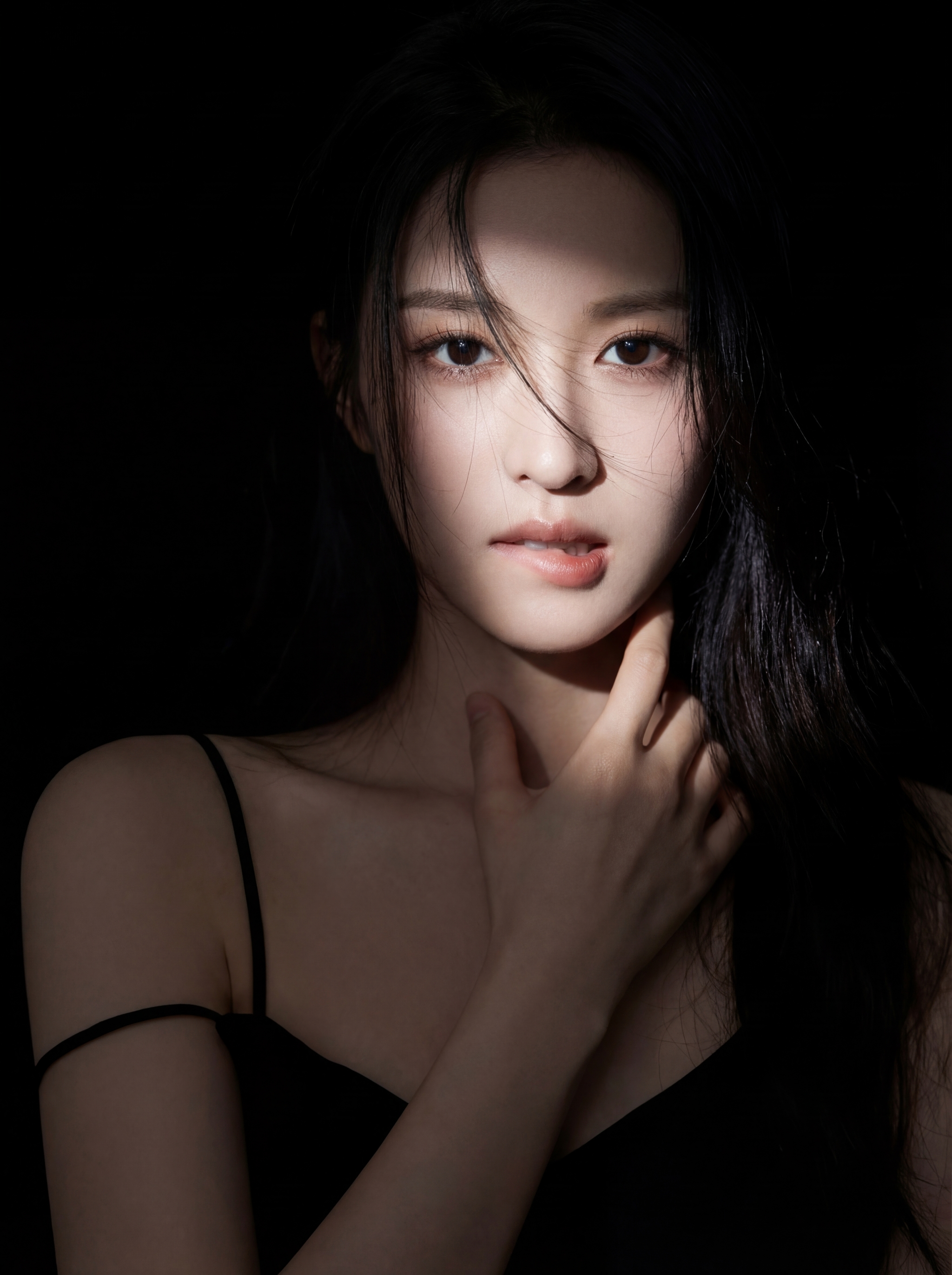 Young woman in a dark setting with a narrow beam of light illuminating her face as she looks at the camera and touches her neck