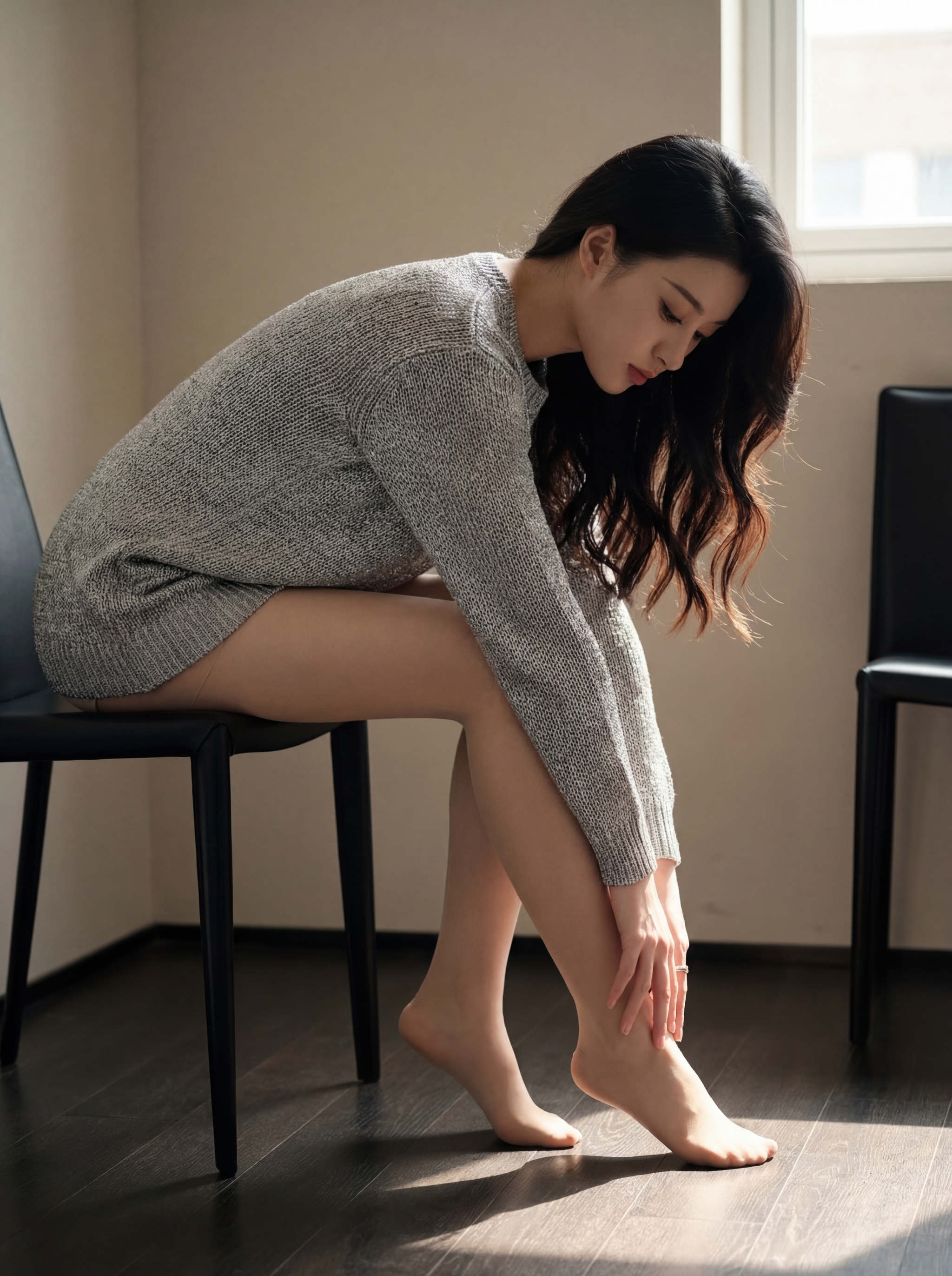 Blurred face woman in gray sweater dress sitting on a chair and reaching toward her bare feet in a sunlit room