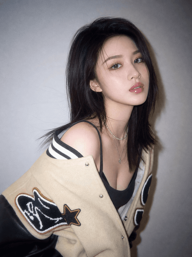 Close up portrait of a person wearing a varsity jacket draped off one shoulder with striped bra straps and a choker necklace against a plain backdrop