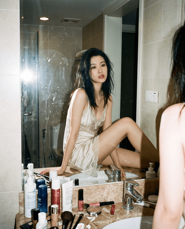 Person sitting on a bathroom counter facing a mirror surrounded by makeup and toiletries