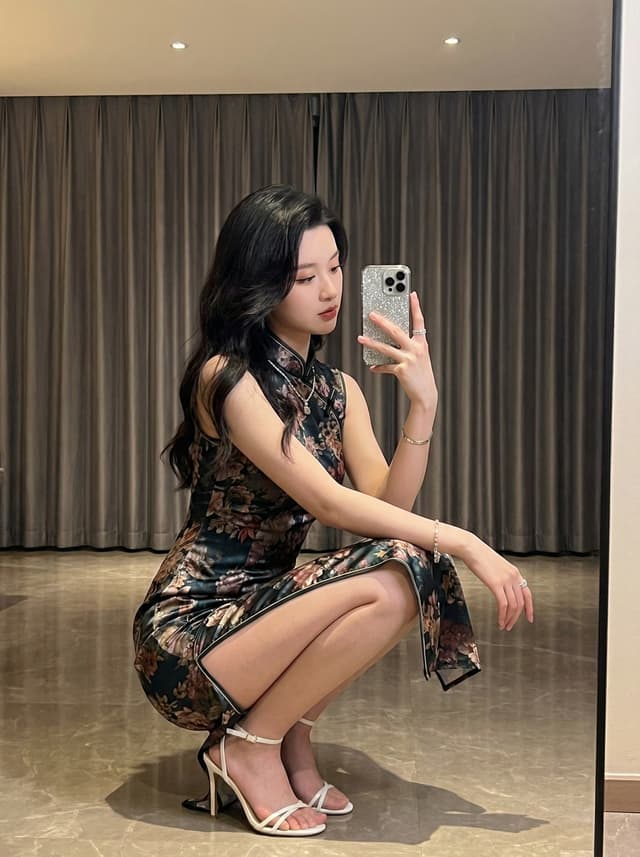 woman in floral dress crouching in high heels taking a mirror selfie in a modern room with floor length curtains